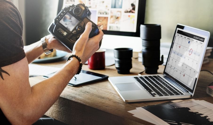 shot of a photographer holding their camera facing their work desk containing a desktop monitor and a laptop