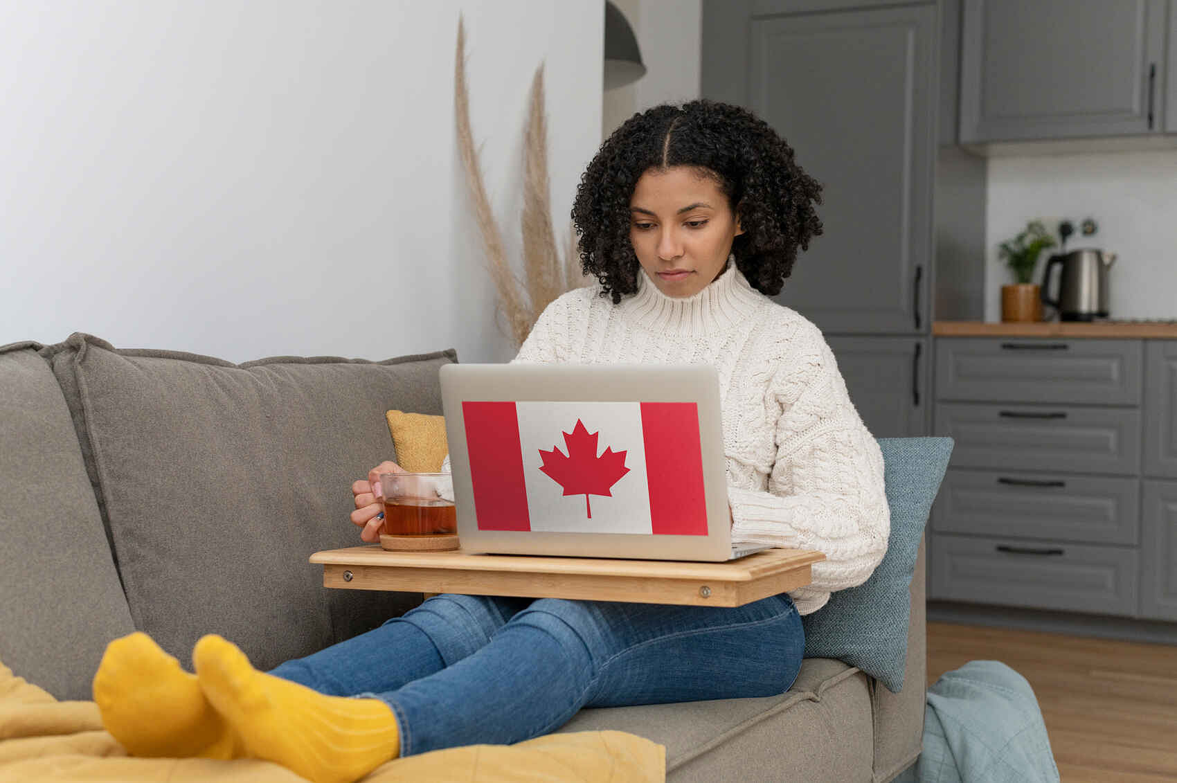 A woman sitting on a couch, working on a laptop adorned with a Canadian flag sticker