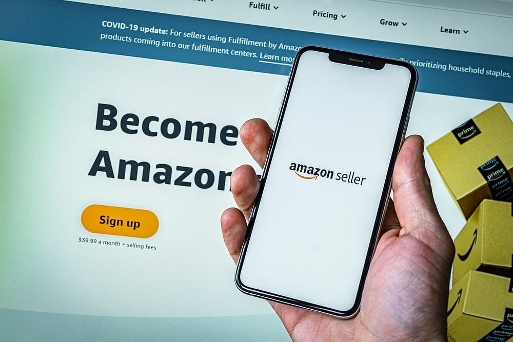 A man holding a smartphone showing the Amazon Seller app, his desktop on the background showing the Amazon Seller Central page