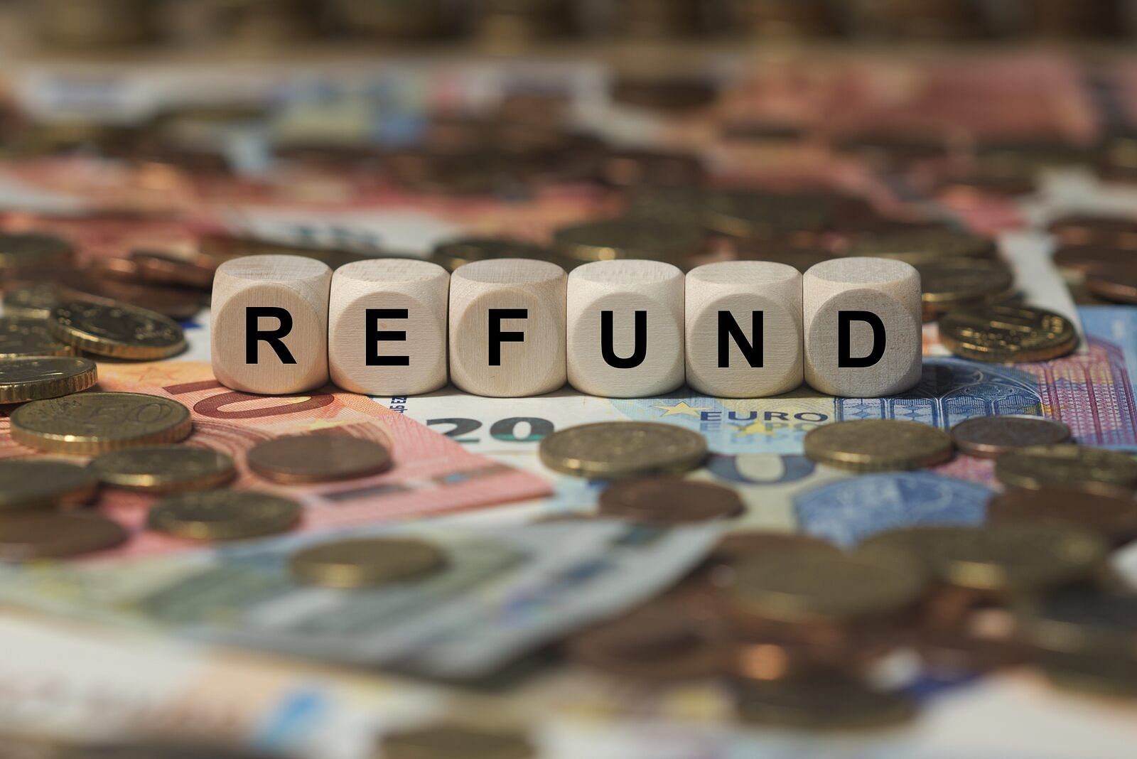 Wooden cubes on top of paper bills and scattered coins are lined up in a row, each letter printed on the cubes spells the word “refund”
