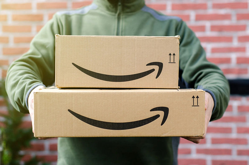 A delivery guy wearing an olive zip-up jacket holds two Amazon boxes with the A-Z logo in front of them; the box at the bottom is wider than the one on top. 
