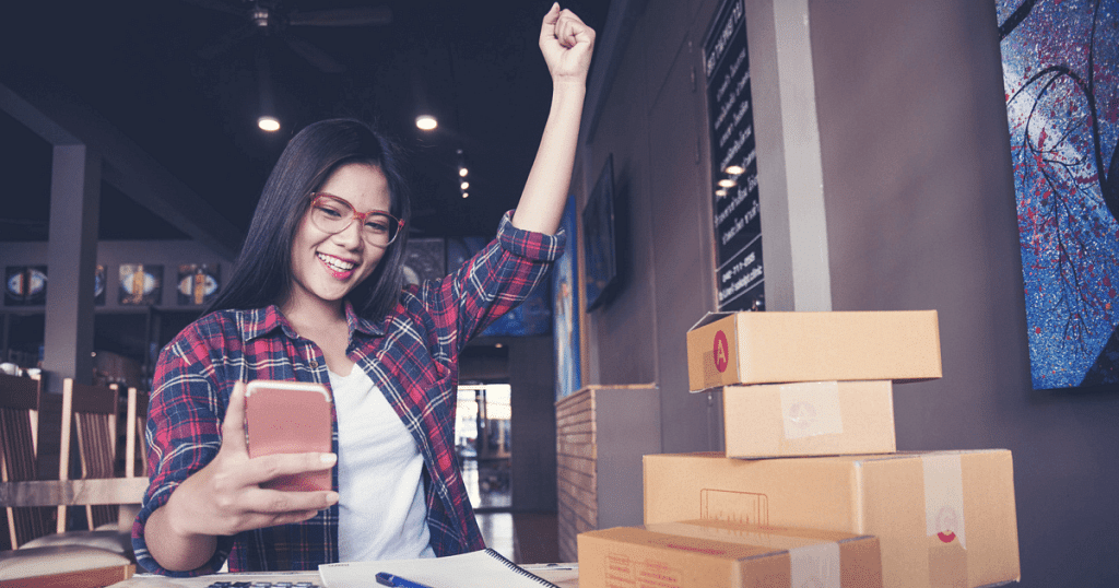 a woman sitting at a table with a notebook and some packages, expressing joy while looking at something on her phone