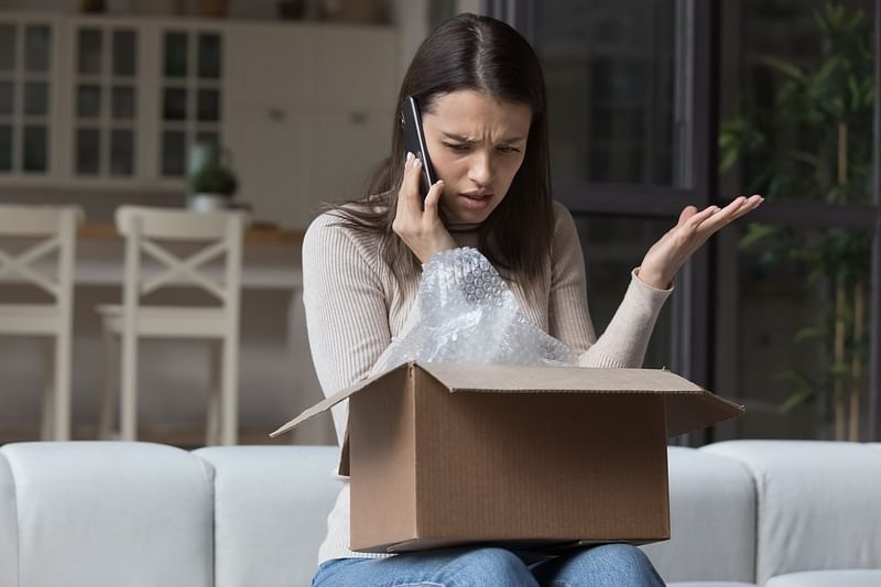 A young woman sits on a couch with an open parcel on her lap complains about the item she received to someone on the other end of her mobile phone.