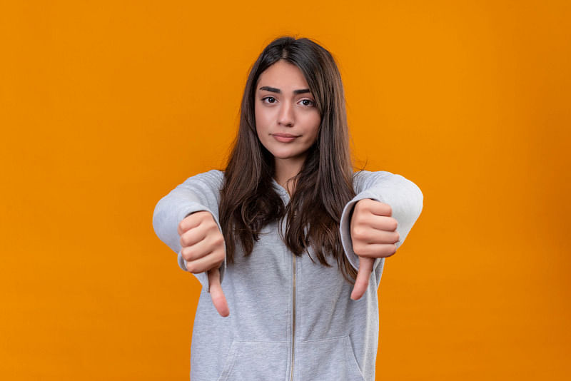 young girl in a gray hoodie looking at the camera, standing in front of an orange background, making the thumbs down gesture with both hands