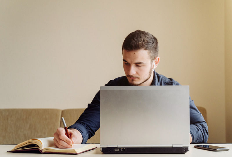 young white man sitting in front of his laptop, writing on his notebook, taking an amazon fba course