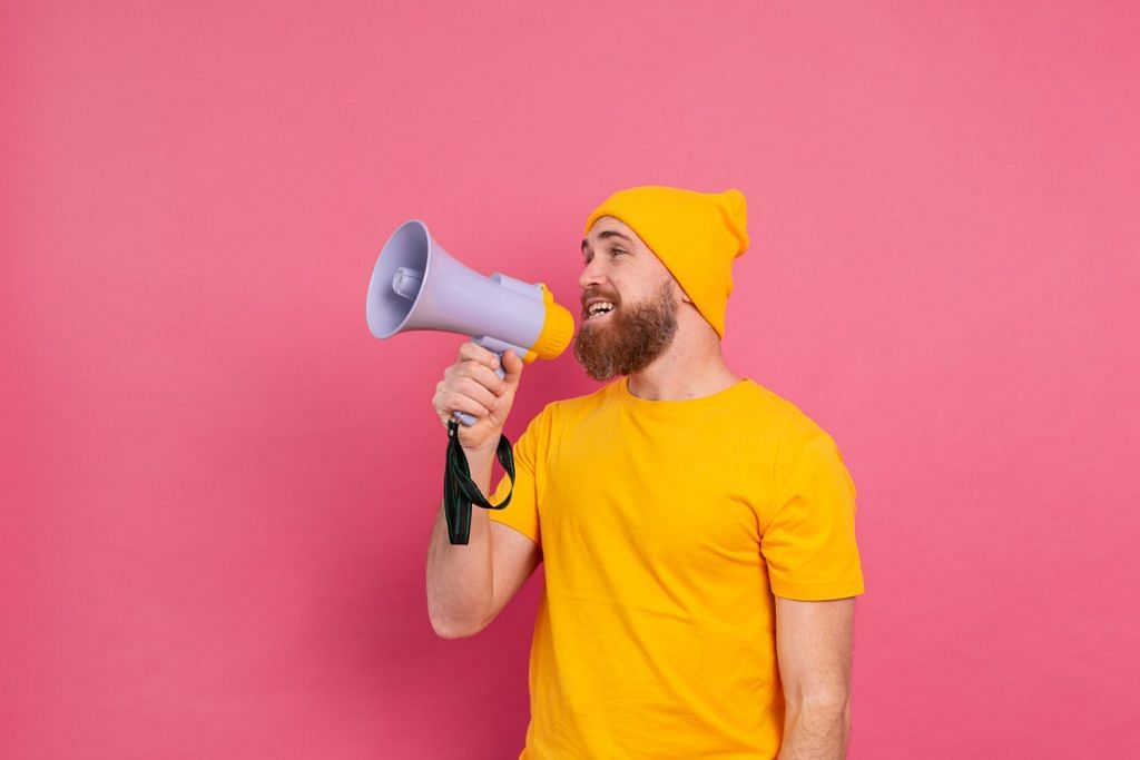 An adult male wearing yellow shirt and beanie holding a megaphone in front of his face